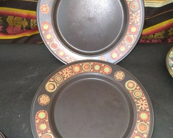 May include: Two brown ceramic dinner plates with a colorful geometric pattern around the rim. The pattern features orange, yellow, green, and white circles and flowers.