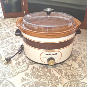 May include: A brown and cream slow cooker with a clear lid. The appliance has a gold-coloured knob and the words "Simmer-Pot" are printed on the front. The slow cooker is plugged into an electrical outlet.