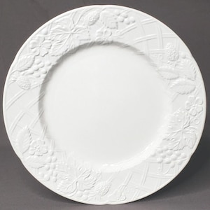 May include: White ceramic dinner plate with embossed floral and fruit design. The rim features a repeating pattern of grapes, strawberries, and flowers, set against a crosshatch background. A classic piece for serving meals.