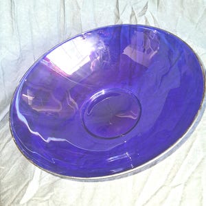 May include: A deep cobalt blue glass bowl with a smooth, rounded interior and a thin, reflective metallic silver rim. The bowl features a wide, open circular shape with a slightly undulating edge, catching light to create bright white reflections.