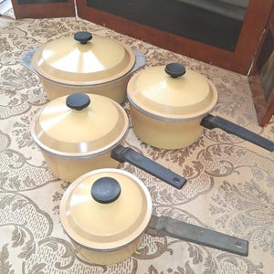Peut inclure: Ensemble de quatre casseroles et poêles vintage en émail jaune avec des poignées et des couvercles noirs. Les casseroles sont de tailles et de formes différentes, avec une palette de couleurs assortie. L'ensemble est posé sur une nappe à motifs.