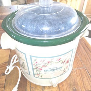 May include: A Rival Crock-Pot slow cooker with a cream-colored base featuring a floral pattern and a dark green rim. It includes a clear glass lid with a central handle and a white power cord. The front control dial is set to off.