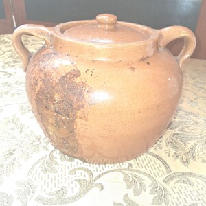 May include: A brown ceramic pot with a lid and two handles. The pot has a rounded shape and a slightly textured surface. The lid has a small knob on top. The pot is sitting on a patterned tablecloth.