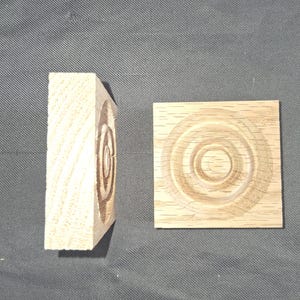 May include: Two square light-coloured wooden blocks featuring a carved concentric circle pattern on the front face. One block is positioned flat to show the full design, while the other is turned on its side to display the thickness and grain.