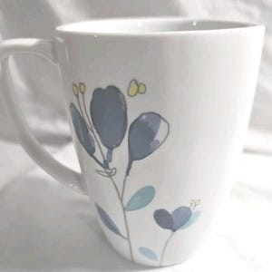 May include: White ceramic mug with a curved handle, decorated with a floral design. The flowers are primarily blue and gray, with accents of yellow and teal. The mug has a smooth, glossy finish and is suitable for hot or cold beverages.