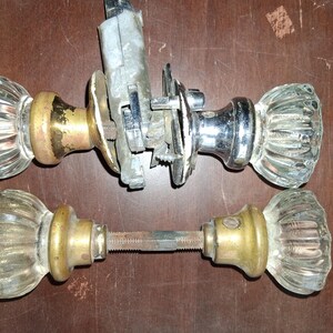 May include: Two sets of vintage door knobs featuring clear, fluted glass handles. One set includes a brass-toned base and a chrome-plated mechanism with mounting hardware. The second set features brass-toned bases connected by a threaded metal spindle.
