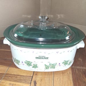 May include: An oval Rival Crock-Pot slow cooker with a white base featuring a green ivy leaf and berry pattern. It has a dark green rim, white side handles, and a clear glass lid with a central knob. The front displays the text Rival Crock-Pot.