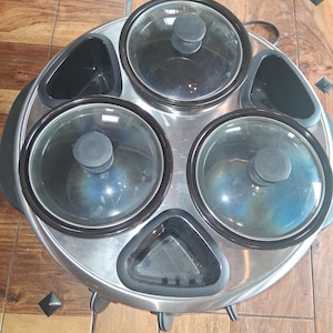 May include: A stainless steel buffet server with four round, glass-lidded warming compartments and three black serving trays. The server has a circular design with a black handle and sits on small legs. Ideal for keeping food warm at gatherings.