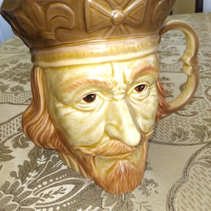 May include: A ceramic pitcher shaped like a man's head with a brown hat and a beard. The pitcher has a cross design on the hat and a handle on the side.