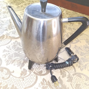 May include: A vintage stainless steel electric coffee pot with a black handle and lid knob. The coffee pot has a spout and sits on a black base. A black power cord with a plug is connected to the pot.