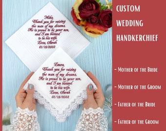 May include: Two white handkerchiefs with lace trim and red embroidered text. The text reads "Mike, Thank you for raising the man of my dreams. He is proud to be your son, and I am blessed to be his wife. Love, Sarah 01/15/2022" and "Laura, Thank you for raising the man of my dreams. He is proud to be your son, and I am blessed to be his wife. Love, Sarah 01/15/2022". The handkerchiefs are held by hands wearing white lace gloves. The background is a light blue wooden surface.