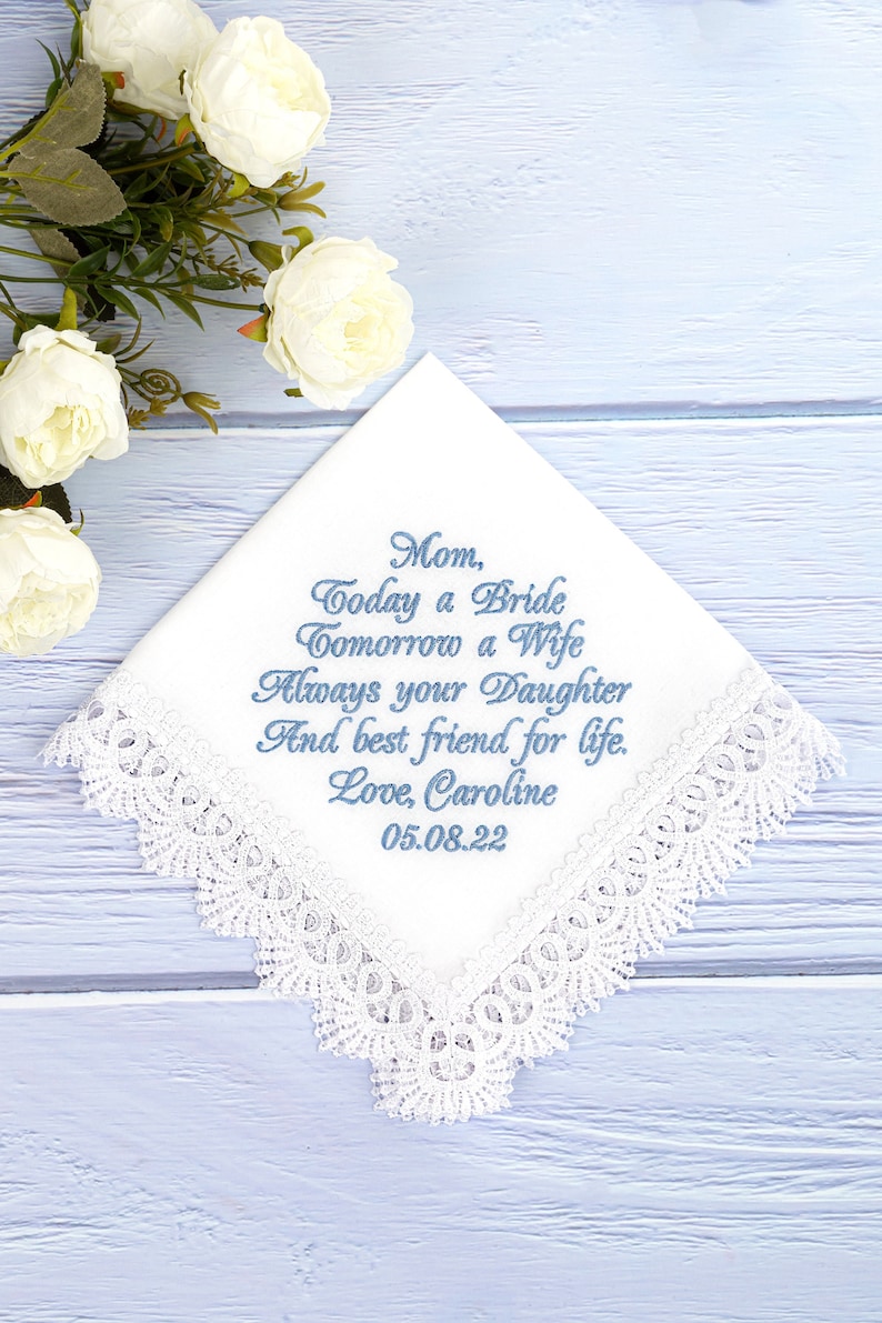 May include: White lace handkerchief with blue embroidered text that reads "Mom, Today a Bride Tomorrow a Wife Always your Daughter And best friend for life. Love, Caroline 05.08.22"