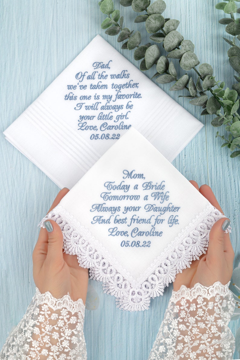 May include: Two white handkerchiefs with lace trim and blue embroidered messages for a bride's father and mother. The father's handkerchief reads "Dad, Of all the walks we've taken together, this one is my favorite. I will always be your little girl. Love, Caroline 05.08.22". The mother's handkerchief reads "Mom, Today a Bride Tomorrow a Wife Always your Daughter And best friend for life. Love, Caroline 05.08.22".