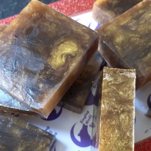 May include: A close-up of several bars of handmade soap. The soap is brown with gold flecks and swirls. The soap is on a white surface with a purple logo that says "Singing Wolf Soap".