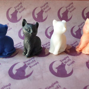 May include: Four small cat-shaped soap figurines in different colours: blue, black, white, and orange. The soaps are arranged in a row on a white and purple patterned surface.