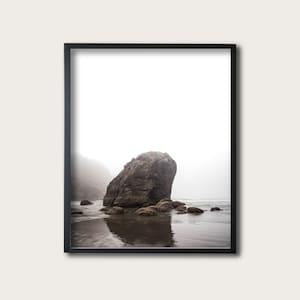 May include: A large, gray rock sits on a beach with a misty background. The rock is partially submerged in the water, and its reflection is visible in the water.