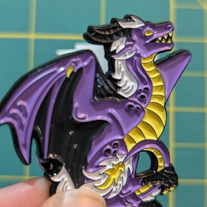 May include: Purple and yellow enamel pin of a dragon with wings spread out. The dragon has a white mane and a black face.