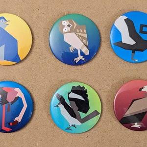 May include: Six colorful round buttons with different geometric animal designs. The buttons feature a penguin, an owl, a swan, a flamingo, a bird, and a vulture.