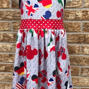May include: A gray sundress featuring Mickey Mouse-shaped designs with flags from various countries. The dress has a red polka-dot waistband and navy blue straps. The flags include the USA, Germany, and the UK.