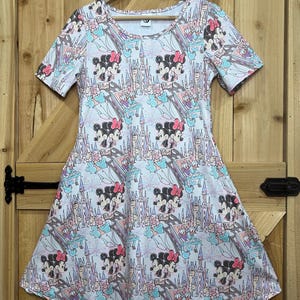 Op de afbeelding: Een witte jurk met een patroon van Minnie Mouse, kastelen en regenbogen. De jurk heeft korte mouwen en een uitlopende rok. Het ontwerp bevat roze, blauwe en paarse accenten, waardoor een grillige en kleurrijke look ontstaat. De jurk hangt aan een houten kleerhanger.