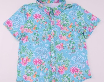 May include: A short-sleeved button-down shirt with a light blue background. The shirt features a floral pattern with pink, green, and orange flowers, along with cartoon character designs and castle illustrations. The shirt has a classic collar.
