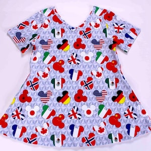 Puede incluir: Vestido azul claro de manga corta con un estampado de cabezas de Mickey Mouse con banderas de varios países. Las banderas incluyen las de EE. UU., Canadá, México y Japón. El vestido tiene un escote redondo y una falda acampanada.