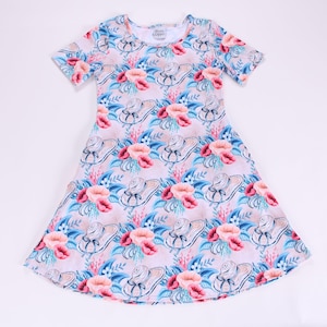 Puede incluir: Vestido de manga corta con estampado floral y de sombrero. El vestido presenta un fondo rosa claro con imágenes repetidas de sombreros, flores rosas y hojas azules. El vestido tiene un cuello redondo.