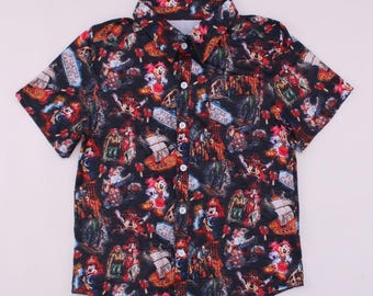 May include: A short-sleeved button-down shirt with a dark background and a colorful pattern featuring cartoon characters, pirate ships, and the text "Pirates Caribbean". The shirt has a chest pocket and white buttons.