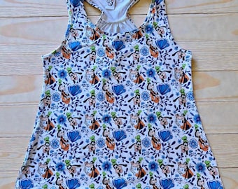 May include: White tank top with a pattern featuring cartoon characters, blue flowers, and green leaves. The tank top has a racerback design and a scoop neckline. The pattern includes orange, blue, and green accents.