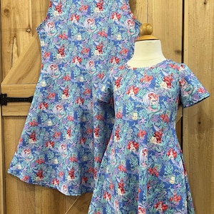 May include: Two blue dresses with a mermaid print. One is a sleeveless dress, and the other has short sleeves. The dresses feature a pattern of mermaids, coral, and seaweed in shades of red, pink, and green.