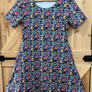 May include: A blue short-sleeved dress with a print of the Hogwarts house crests. The crests are in their traditional colours: Gryffindor (red and gold), Hufflepuff (yellow and black), Ravenclaw (blue and bronze), and Slytherin (green and silver).