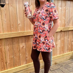 May include: A woman wearing a tunic and leggings with a fall-themed print, holding a tumbler. The tunic features pumpkins, sunflowers, and coffee cups in shades of orange, pink, and brown. She is also wearing brown leggings and sandals.