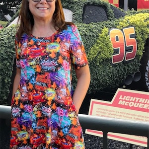 May include: A woman wearing a colorful dress featuring Cars movie characters. She is also wearing a black and white checkered bow headband and sunglasses. The number 95 is visible in the background.