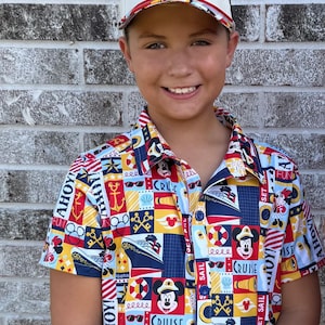 May include: A white baseball cap with a gold embroidered key emblem and a short-sleeved button-down shirt featuring a colorful nautical-themed print with cruise ships, anchors, life preservers, and cartoon characters with the text AHOY and CRUISE.