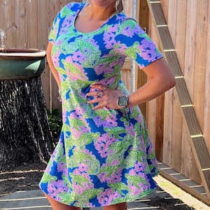 Puede incluir: Vestido de manga corta con fondo azul y estampado de tortugas verdes y coral rosa. El vestido llega a la rodilla y tiene cuello redondo. La persona lleva sandalias tejidas.
