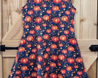 May include: A sleeveless dress with a navy blue backdrop, adorned with a repeating pattern of orange pumpkins and autumnal leaves. The dress is displayed on a wooden hanger, set against a wooden door. It has a scoop neckline and a flared skirt.