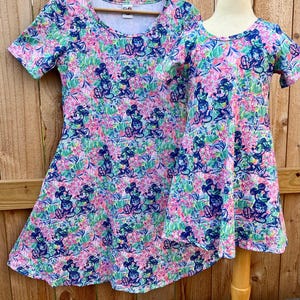 May include: Two dresses with a colorful floral and Mickey Mouse print. The dresses have short sleeves and a scoop neckline. One dress is on a wooden hanger, and the other is on a dress form. The background is a wooden fence.