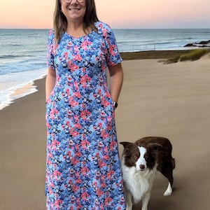May include: A woman wearing a blue and pink floral maxi dress with pockets stands on a sandy beach with a brown and white border collie dog.