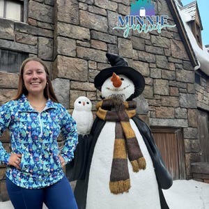 May include: A woman wearing a blue and white patterned shirt with a zipper and a pair of dark blue leggings stands in front of a snowman wearing a black hat and a brown and yellow scarf. The snowman is standing in front of a stone building with a sign that reads "MINNIE jewels".