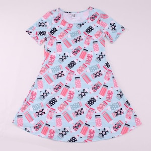 Puede incluir: Vestido azul claro de manga corta con cuello redondo. El vestido presenta un estampado repetido de lápices rosas, negros y florales. Hecho de una tela suave y fluida, perfecto para el uso diario.