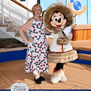 May include: A woman in a white dress with a black and red Minnie Mouse pattern poses with Minnie Mouse, who is dressed in a winter outfit. Minnie Mouse wears a white coat with fur trim and a fur-lined hood. The background shows a wooden deck and a blue sky.