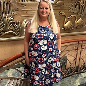 May include: A woman in a navy dress patterned with anchors, life rings, and ships, wearing Mickey Mouse ears with a red bow. The background is a gold-toned wall with a relief of a landscape. The dress has a knee-length hem.