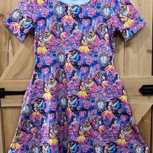 May include: A short-sleeved dress with a vibrant, all-over print featuring characters and elements from a fairy tale. The design includes images of a princess, a beast, clocks, roses, and hearts in shades of pink, purple, and blue.