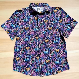 May include: Short-sleeved button-down shirt with a colorful pattern featuring Mickey Mouse jack-o'-lanterns, ghosts, and ornate floral designs. The shirt's background is a dark blue, with accents of purple, pink, yellow, and green.