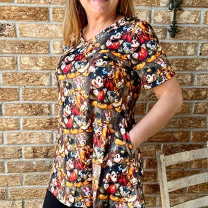 May include: A short-sleeved tunic top with a colourful print of Mickey Mouse and friends. The top is worn with black shorts. The design features a variety of characters in a palette of red, yellow, and black.