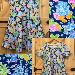 以下が含まれることがあります： ネイビーブルーの花柄のノースリーブドレスと半袖ドレス。ピンク、青、緑、オレンジの花、お城、漫画のキャラクターのデザインが含まれています。ドレスは木製のハンガーに掛けられています。