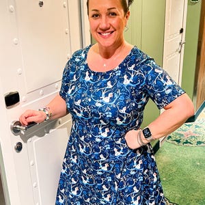 Pode incluir: Uma mulher usa um vestido azul com um padrão branco e azul, mangas curtas e saia rodada. Ela sorri, usa uma faixa branca e joias de prata. A porta atrás dela tem o número 8166. O padrão do vestido é floral.
