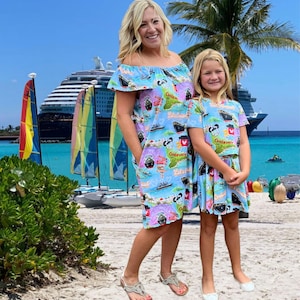 May include: Two matching light blue dresses with a nautical print, featuring cruise ships, maps, and the text "Castaway". A woman and a child wear the dresses on a beach with a cruise ship in the background.