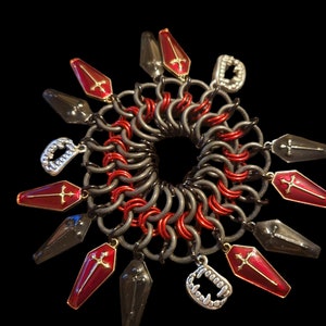 May include: A black and red chainmail bracelet with coffin-shaped charms featuring crosses and fangs. The charms are silver and gold with red enamel.