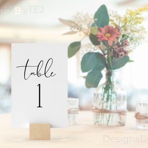 Könnte beinhalten: Weiße Karte mit schwarzem Text, der "Tisch 1" lautet, auf einem Holzständer. Die Karte liegt auf einem Tisch mit einer Blumenvase im Hintergrund.